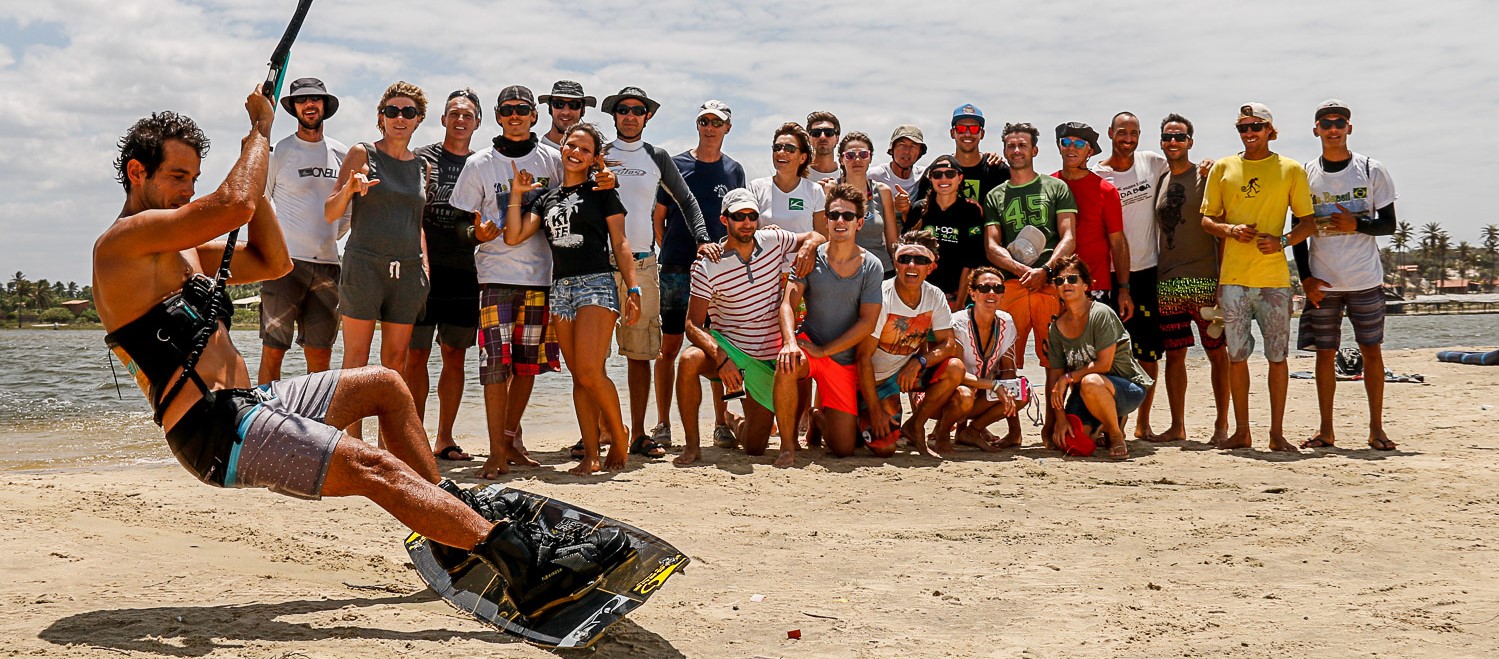 Crowd watches as man rides across sand on heel edge of his kitesurf board.